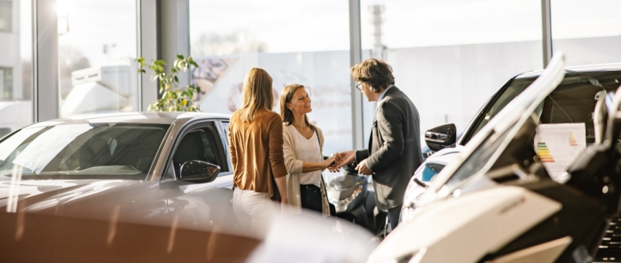 Hombre de ventas saludando la mano de una clienta de pie junto a otra mujer en una sala de exposición de un concesionario de autos