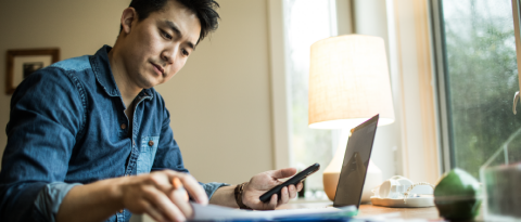 A man at a desk consults a paper and his phone while paying bills